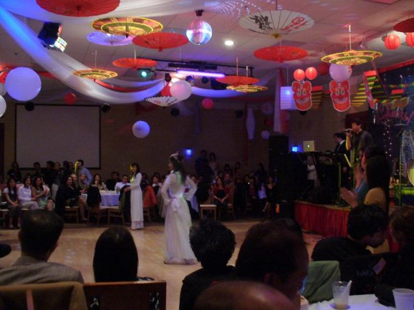 A woman in white traditional Vietnamese dress stands alone on a dance floor as a band performs behind her (right) and an audience looks on (background and foreground).